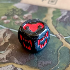 Puede incluir: Un dado negro y rojo con un símbolo de cabeza de carnero rojo en un lado y dientes rojos en los otros lados.