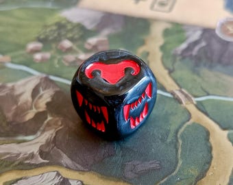 May include: A six-sided gaming die with a glossy black finish and recessed red engravings. The faces feature stylised designs including a horned emblem and sharp, tooth-like patterns carved into the surface of the cube.