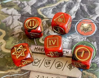May include: Five red and gold dice with Roman numerals and symbols are displayed on a game board. The dice are a marbled red with gold accents. The game board has a map design with text and circular tokens.