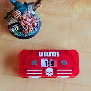 May include: Red plastic counter with a white skull and the word "WOUNDS" printed on it. The counter shows the number "19".
