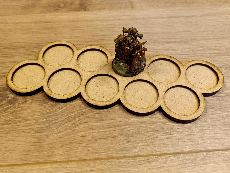 May include: A wooden miniature figure display tray with ten circular cutouts. A painted miniature figure is placed in one of the cutouts. The tray is made of light brown wood and is designed to hold small figurines.