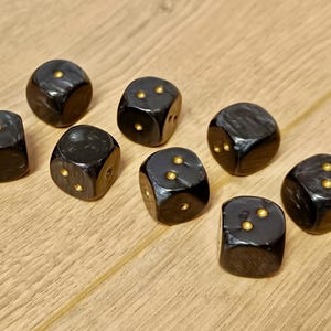 May include: Eight black dice with gold-colored dots are arranged on a light brown wooden surface. The dice are glossy and have rounded edges. The dots are evenly spaced on each face, indicating the numbers.