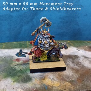 May include: A 50 mm x 50 mm movement tray adapter for Thane & Shieldbearers. The tray features three painted miniatures of dwarves, one with a raised axe, one with a shield, and one with a raised hammer. The tray is made of wood and has a black base.