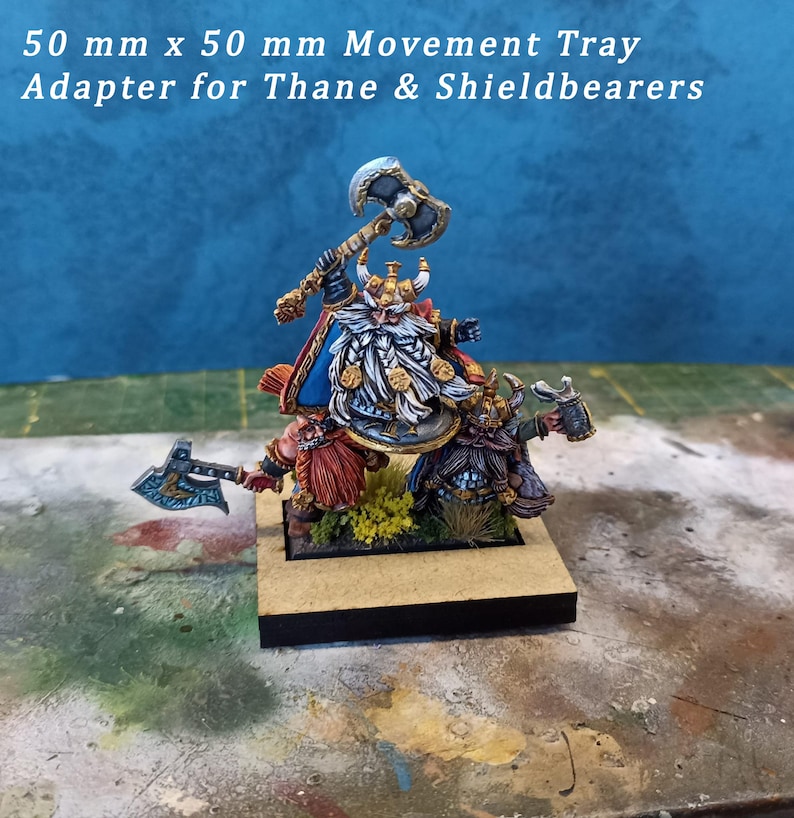 May include: A 50 mm x 50 mm movement tray adapter for Thane & Shieldbearers. The tray features three painted miniatures of dwarves, one with a raised axe, one with a shield, and one with a raised hammer. The tray is made of wood and has a black base.