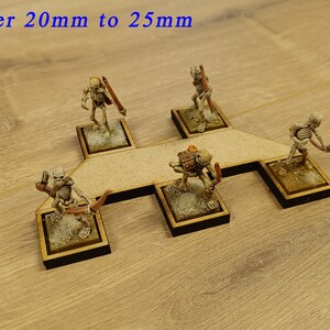 May include: Five miniature skeletal figures with bows and arrows are arranged on a wooden base. The base is shaped like a cross with a central platform. The figures are painted in a light brown colour. The text "Open Order 20mm to 25mm" is visible at the top of the image. The text "Group" is visible at the bottom of the image.