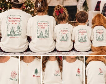 Pode incluir: Um conjunto de camisas cor de creme a combinar e uma camisola para animal de estimação com um gráfico nas costas de uma árvore de Natal, um trenó e o texto The Williams Family Crew 2026 Nashville TN. Os designs frontais incluem ícones como Pai Natal, árvore, laço, boneco de neve e rena.