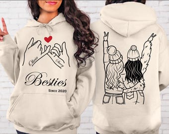 May include: A cream-colored hoodie with a black outline of two girls holding hands with the text "Olivia" and "Stacey" written on their wrists. The text "Besties Since 2020" is printed below the hands. The back of the hoodie features a black outline of two girls with their arms around each other, wearing winter hats.