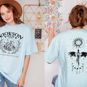 A woman wearing a light blue t-shirt with a graphic design and text, posing in front of a floral wall.