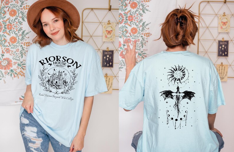 A woman wearing a light blue t-shirt with a graphic design and text, posing in front of a floral wall.