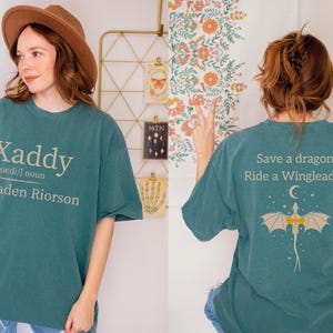 A woman wearing a green t-shirt with a dragon design and text on the back, posing in front of a floral wall.
