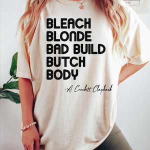 May include: Cream-colored t-shirt with the words "BLEACH BLONDE BAD BUILD BUTCH BODY" in bold black letters. Below the text is "-A Crockett Clapback" in a handwritten style font. The shirt is paired with dark green shorts.