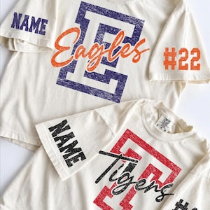May include: Two off-white t-shirts with distressed graphic prints. The top shirt features a purple letter E, orange script text reading Eagles, and sleeve text reading NAME and #22. The bottom shirt has a red letter T, black script text reading Tigers, and sleeve text reading NAME and #18.
