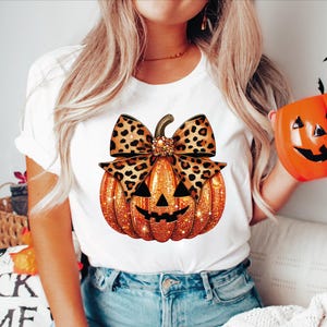May include: White t-shirt featuring a glittery orange pumpkin with a carved face and a leopard print bow. The pumpkin design is centered on the shirt, perfect for Halloween. The shirt is paired with blue jeans.