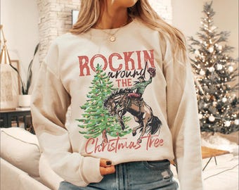 May include: Cream-colored sweatshirt featuring a festive design. The graphic includes a Christmas tree, a cowboy riding a horse, and the text "ROCKIN' around the Christmas Tree" in red and green. The sweatshirt is a long-sleeved crew neck.