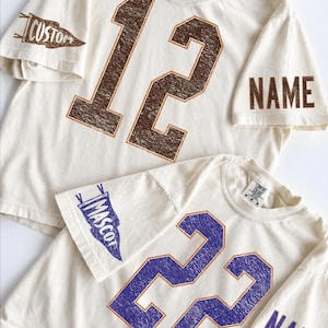 May include: Two off-white t-shirts featuring distressed-style numbers on the front. The top shirt has the number 12 in brown, a pennant graphic with the word CUSTOM on one sleeve, and NAME on the other. The bottom shirt has the number 22 in purple, a pennant graphic with the word MASCOT on one sleeve, and NAME on the other.