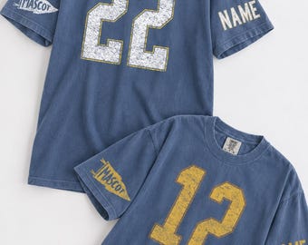 May include: Two blue short-sleeve t-shirts with a distressed finish. One shirt features the number 22 in white, the other the number 12 in yellow. Both shirts have the word MASCOT on the right sleeve and the word NAME on the left sleeve.
