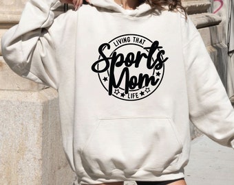 May include: Cream-colored hoodie with a front pocket and drawstring hood. The black graphic on the front reads "LIVING THAT Sports Mom LIFE" in a circular design with stars. The hoodie is a casual, comfortable garment.