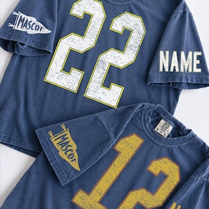 May include: Two blue short-sleeve t-shirts with distressed graphics. The top shirt features the number 22 in white with yellow outlines, a pennant graphic on the sleeve with the text MASCOT, and the word NAME on the other sleeve. The bottom shirt features the number 12 in yellow, a pennant graphic with MASCOT, and the word NAME.