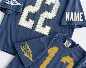 May include: Two blue short-sleeve t-shirts with distressed graphics. The top shirt features the number 22 in white with yellow outlines, a pennant graphic on the sleeve with the text MASCOT, and the word NAME on the other sleeve. The bottom shirt features the number 12 in yellow, a pennant graphic with MASCOT, and the word NAME.