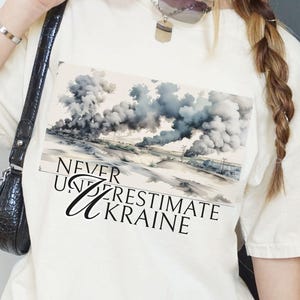 May include: Cream-coloured t-shirt featuring a landscape graphic with smoke and the text "Never Underestimate Ukraine" in black. A small black camera is visible on the front of the shirt.