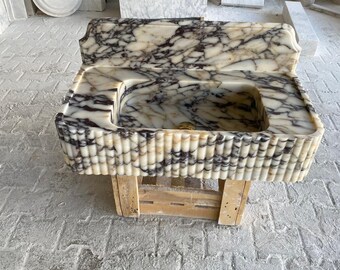May include: A white marble sink with a decorative, fluted edge. The sink is mounted on a wooden stand and has a rectangular shape.