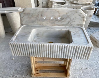 May include: White marble sink with a fluted edge and a backsplash. The sink is mounted on a wooden stand.