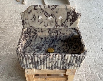 Puede incluir: Un lavabo de mármol blanco y morado con un borde decorativo acanalado. El lavabo tiene una forma rectangular y está montado en una tarima de madera.