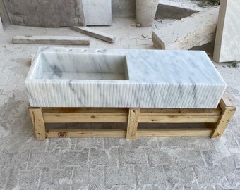 Puede incluir: Un lavabo de baño de mármol blanco con una cuenca rectangular y un borde acanalado. El lavabo está montado en un marco de madera.