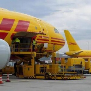 May include: A yellow and red DHL cargo plane with its cargo door open and a yellow loading ramp attached. The plane is parked on the tarmac with another DHL plane in the background.