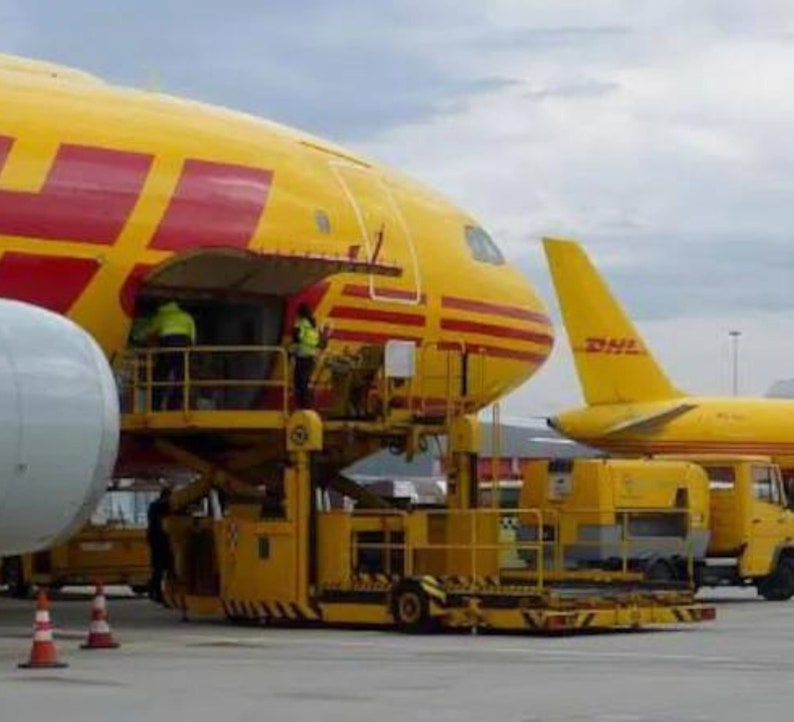 May include: A yellow and red DHL cargo plane with its cargo door open and a yellow loading ramp attached. The plane is parked on the tarmac with another DHL plane in the background.
