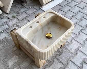 Puede incluir: Un lavabo de mármol blanco con un borde acanalado, colocado sobre un soporte de madera. El lavabo tiene tres agujeros para grifos y un tapón de desagüe dorado.