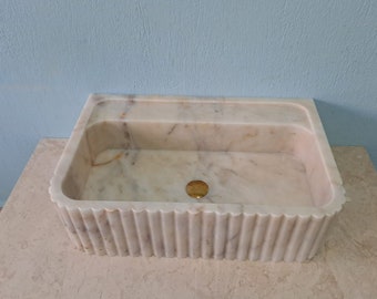 May include: A white marble sink with a fluted edge and a single drain hole. The sink is set on a beige countertop.
