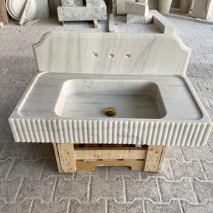 May include: White marble bathroom sink with a backsplash and a fluted edge. The sink is mounted on a wooden pallet.