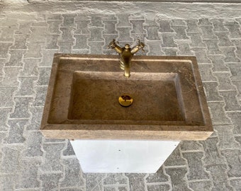 Puede incluir: Un lavabo de piedra marrón con un grifo y un desagüe dorados. El lavabo está montado sobre un pedestal blanco.