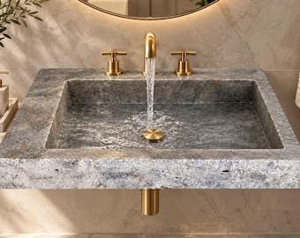 Puede incluir: Un lavabo rectangular de piedra gris con un grifo de oro cepillado y dos mandos en cruz a juego. El agua fluye desde el caño central hacia la cubeta, que cuenta con una tapa de desagüe dorada y un tubo dorado visible debajo.