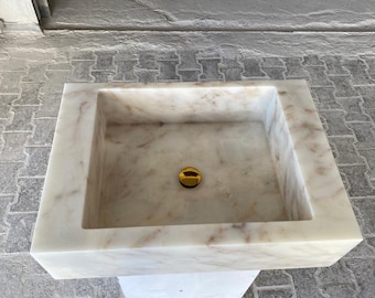 Puede incluir: Un lavabo de baño de mármol blanco con un tapón de desagüe dorado. El lavabo es cuadrado y tiene un borde elevado.