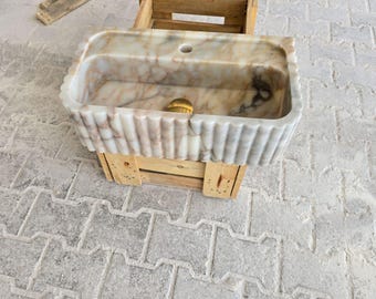 Op de afbeelding: Een rechthoekige marmeren wastafel met een geschulpte rand, met beige, bruine en witte tinten. De wastafel staat op een houten krat, wat een natuurlijk stenen ontwerp benadrukt. Een klein, rond, goudkleurig object ligt erin.