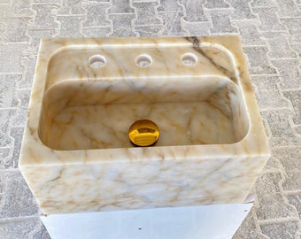 Puede incluir: Un lavabo de mármol blanco con un tapón de desagüe dorado. El lavabo tiene tres agujeros para grifos.