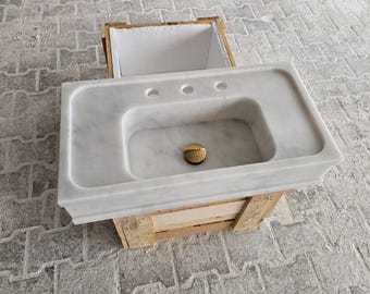 May include: A white marble bathroom sink with a rectangular basin and three tap holes. The sink has a gold-coloured drain and is set in a wooden crate. The sink is made of white marble.