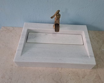 May include: White marble bathroom sink with a brass faucet. The sink has a rectangular shape and a built-in soap dish.