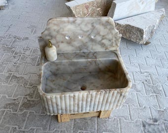 May include: A white marble sink with a decorative fluted edge, mounted on a wooden stand. The sink has a soap dispenser and a drain.