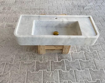 Puede incluir: Un lavabo de baño de mármol blanco con forma rectangular y un borde acanalado. El lavabo está montado sobre un soporte de madera.
