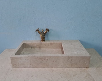 May include: A rectangular, light beige marble sink with a single brass faucet. The sink is set on a matching marble countertop.