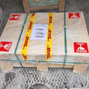 May include: A wooden crate with a yellow DHL tape and a red label that says "DİKKAT KIRILIR FRAGILE" in Turkish and English. The crate is wrapped in clear plastic.