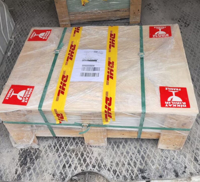 May include: A wooden crate with a yellow DHL tape and a red label that says "DİKKAT KIRILIR FRAGILE" in Turkish and English. The crate is wrapped in clear plastic.