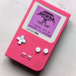 May include: A pink Game Boy Pocket handheld console. The screen shows a purple Mega Man game. The console has white buttons and a directional pad. The text "GAME START PASS WORD" and "LICENSED TO NINTENDO" are visible.