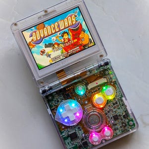 May include: A transparent Game Boy Advance SP console showing the game "Advance Wars". The console's internal workings are visible, with multi-coloured illuminated buttons. The screen displays the text "Press Start!"