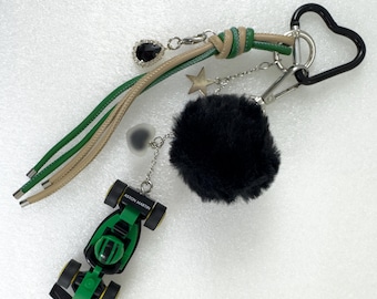 May include: A keychain with multiple charms. It features a green and tan leather strap, a black heart-shaped clasp, a black pom-pom, a star charm, and a small green race car toy with the text "ASTON MARTIN". Additional charms include a black gem and a heart-shaped charm.