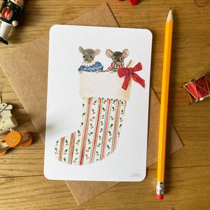 Puede incluir: Una tarjeta de Navidad con dos ratones mirando por encima de un calcet&iacute;n festivo. El calcet&iacute;n tiene rayas rojas y verdes y acebo. Un lazo rojo est&aacute; atado en la parte superior. Un tren de madera rojo y otras decoraciones est&aacute;n en el fondo.