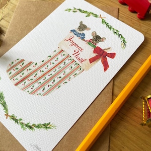 Puede incluir: Tarjeta de Navidad con una ilustraci&oacute;n en acuarela de un calcet&iacute;n y dos ratones. El calcet&iacute;n tiene rayas y bayas rojas y verdes, con las palabras "Joyeux No&euml;l". La tarjeta est&aacute; sobre un sobre marr&oacute;n.
